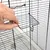 Main ouvrant une petite porte en métal sur la cage à barreaux noirs, détail du système de fermeture visible.