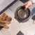 Hand hält einen schwarzen Futternapf mit Marmormuster und Trockenfutter. Hund schaut auf den Napf.