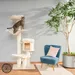 Arbre à chat en bois clair avec griffoirs, plateformes et coussins crème, un chat tigré dessus. Fauteuil bleu, coussin rose et plante verte visibles à côté.