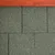 Detalle de tejas asfálticas verdes con borde superior de madera visible. Material rugoso y rectangular, adecuado para techos de casetas u otras estructuras exteriores.
