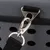 Gros plan sur une attache métallique avec mousqueton et sangle noire fixée à un sac en tissu gris à trous d’aération et fermeture éclair visible.