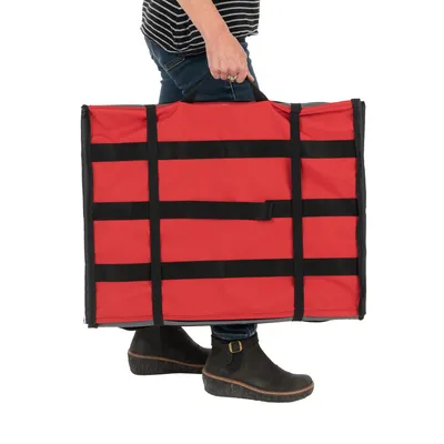 Sac de transport rectangulaire rouge avec poignées noires, porté à la main par une personne en jean et bottines noires. Matière textile visible.
