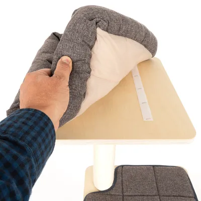 Hand hebt ein graues, gepolstertes Kissen von einer Holzplattform mit Klettverschluss. Darunter eine weitere gepolsterte Matte sichtbar.