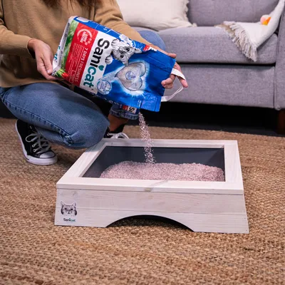 Person pouring Sanicat litter from a bag into a rectangular tray, demonstrating how to fill the tray with litter.