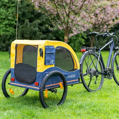 Yellow-blue bicycle trailer with mesh windows and the inscription 'Hafenbande', attached to a black bicycle, on a lawn outdoors.