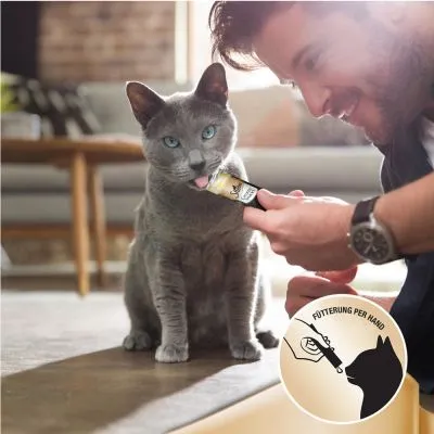 Kissa saa tahnamaista herkkua suoraan kädestä. Alakulmassa kuvake ja teksti: Fütterung per Hand, jossa käsi syöttää kissalle tuotetta.