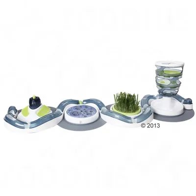 Jouet interactif pour chat composé de plusieurs modules reliés : brosse, plateau à trous, herbe à chat et tour transparente avec balles vertes. Matériaux visibles : plastique, poils synthétiques.