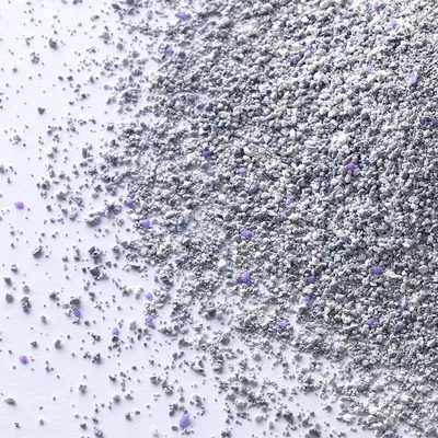 Arena para gatos de grano fino en tonos grises con pequeños gránulos morados visibles, extendida sobre una superficie blanca.