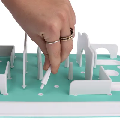 A hand shows a white stick being removed from a green interactive toy. Toy with obstacles and openings for activities.