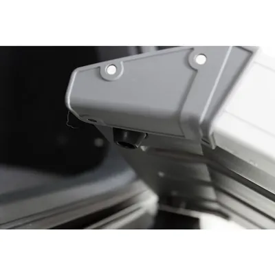 Zbliżenie na plastikowy narożnik szarej skrzynki lub transportera z widocznymi śrubami mocującymi i otworem wentylacyjnym u dołu.