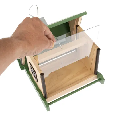 Una mano mostra l'apertura di una casetta per uccelli in legno verde con frontale in plexiglas e gancio in metallo per appenderla.
