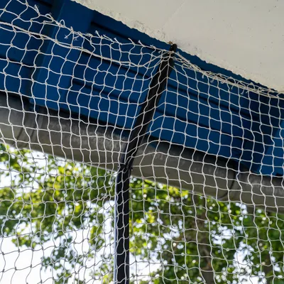 Detailný záber na bielu ochrannú sieť pripevnenú na modrej kovovej konštrukcii a betónovom strope, v pozadí sú viditeľné stromy.