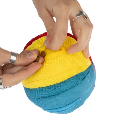 Main insérant des croquettes dans une balle en tissu multicolore jaune, bleu et rouge, ouverture visible sur le dessus de la balle.