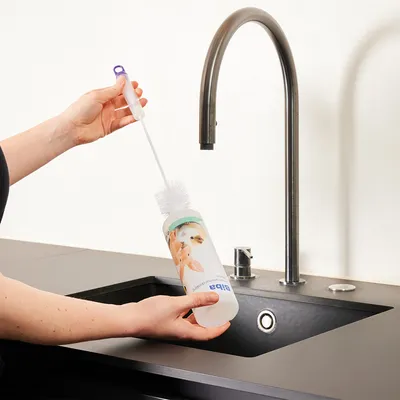 Egy kéz kefével tisztít egy ivópalackot csap alatt. A palackon kisállatok képe és a 'Biba' felirat látható.