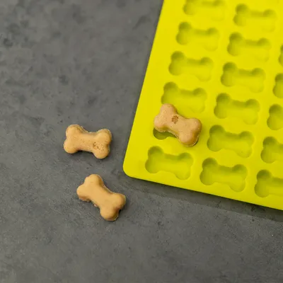 Gul silikoneform med små udtagne hundekiks i benform på grå overflade. Formen har flere fordybninger til bagning af godbidder.