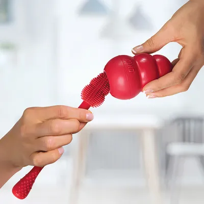 Piros KONG játék tisztító kefével egy piros KONG játékba helyezve, tisztítási használatot mutat.