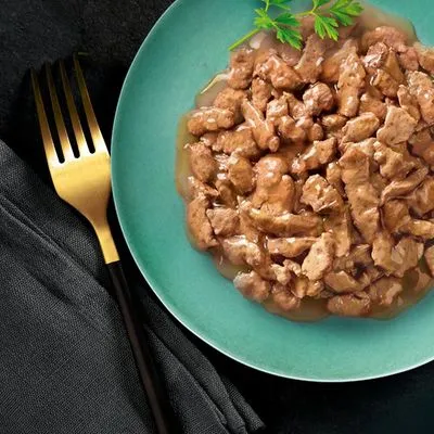 Bord met stukjes natvoer in saus, geserveerd op een groen bord met peterselie als garnering. Gouden vork en zwarte servet zichtbaar naast het bord.