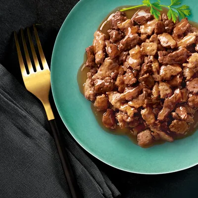 Close-up of juicy meat pieces in sauce on a green plate, garnished with parsley. Golden fork and black cloth next to it.