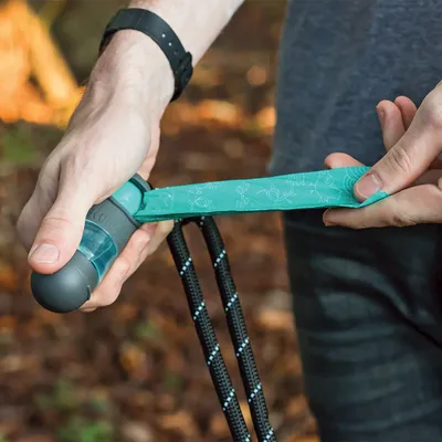 Persoon toont het gebruik van een hondenpoepzakjesdispenser met groene zak, zwarte lijn, grijze handgreep en blauwe deksel.