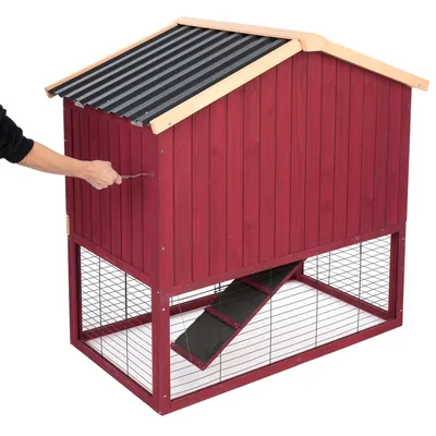 Rotes Holzgehege mit schwarzem Dach, unterer Bereich aus Metallgitter. Hand öffnet obere Tür mit Metallgriff. Rampe verbindet oberen und unteren Bereich.