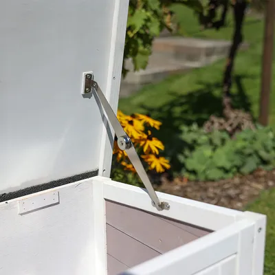 Detailní pohled na bílou dřevěnou nádobu s otevřeným víkem a kovovým držákem. V pozadí zahrada s květinami a rostlinami.