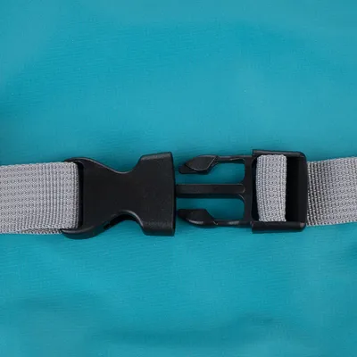 Gros plan sur une sangle grise avec boucle en plastique noir, posée sur un tissu bleu. Boucle ouverte, parties mâle et femelle visibles.