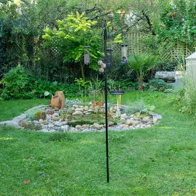 Schwarzer Metallständer mit mehreren Futterstationen für Vögel, darunter Körner- und Meisenknödelhalter, aufgestellt in einem Garten neben einem Teich.