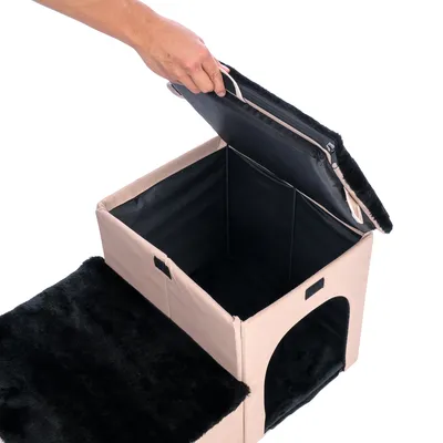 Faltbare beige Haustierhöhle mit schwarzem Plüschbezug, abnehmbarem Deckel und seitlichem Eingang. Hand zeigt Öffnung des Deckels.