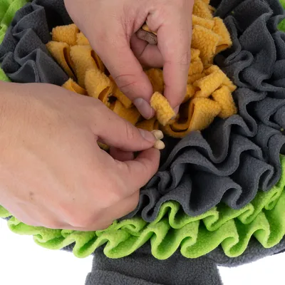 Hände verstecken Leckerlis in einem Schnüffelteppich aus grauem, gelbem und grünem Stoff.