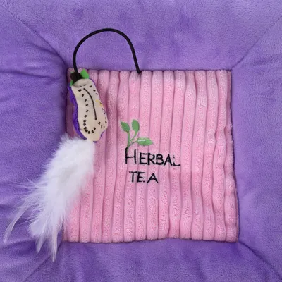 Rosa und lila Stoffspielzeug mit gesticktem Text 'Herbal Tea' und einer weissen Feder an einem schwarzen Band.