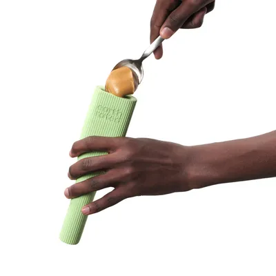 Grüne Silikonhülle mit der Aufschrift 'SodaPup', in die Erdnussbutter mit einem Löffel eingefüllt wird.