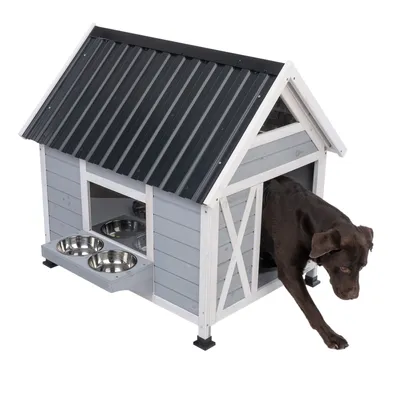 Gråt hundehus med sort tag, integreret fodringsområde og to skåle i rustfrit stål. En brun hund forlader huset gennem døren.