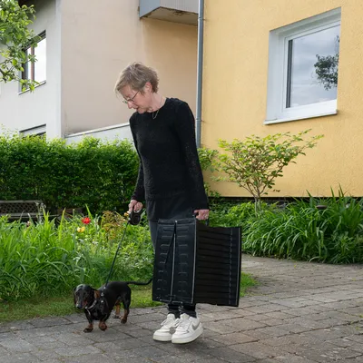 Személy fekete kutyapórázzal és hordozható fekete rámpával, háziállatok számára alkalmas. Környezet: kerti út sárga épület előtt.