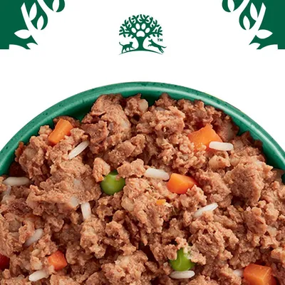Primo piano di cibo umido con pezzi di carne, carote, piselli e riso in una ciotola verde. Logo con albero e animali visibile in alto.