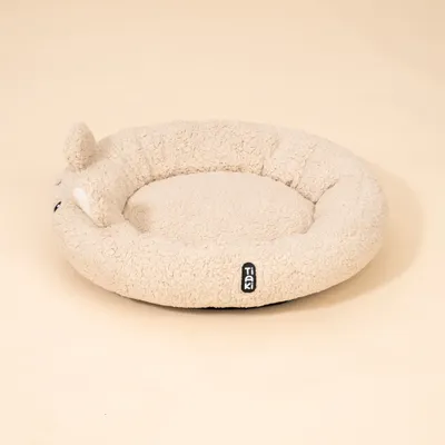 Rundes, beiges Kuschelbett mit Tiaki-Label und Ohren-Applikation aus weichem Stoff. Rundes, beiges Kuschelbett mit Tiaki-Label und Ohren-Applikation aus weichem Stoff.