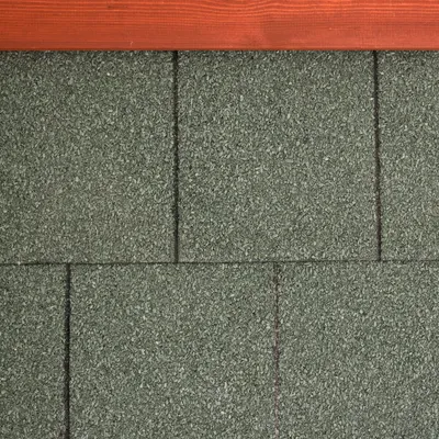 Detalle de tejas asfálticas verdes con borde superior de madera visible. Material rugoso y rectangular, adecuado para techos de casetas u otras estructuras exteriores.