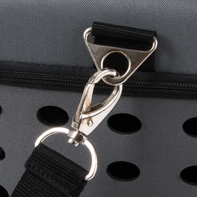 Gros plan sur une attache métallique avec mousqueton et sangle noire fixée à un sac en tissu gris à trous d’aération et fermeture éclair visible.