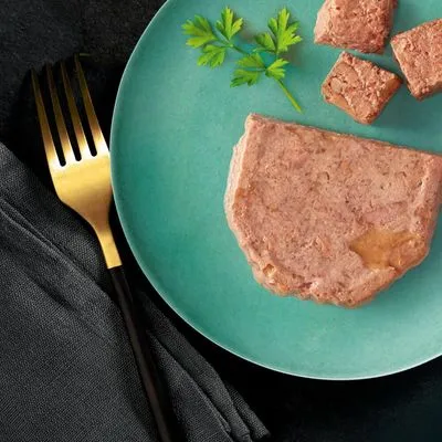 Portie natvoer in blokjes en schijf op groen bord met peterselie, naast gouden vork. Geen merknaam of verpakking zichtbaar.