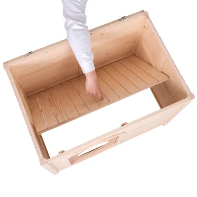 Caisse en bois clair ouverte, une main soulève un plateau intérieur amovible. Poignée découpée visible sur le côté. Matériau : bois.