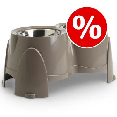Comedero doble elevado de plástico beige con cuencos metálicos. Círculo rojo con símbolo de porcentaje (%) indica oferta o descuento visible en la imagen.