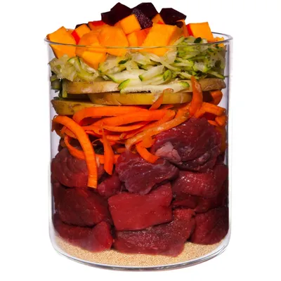 Glass container with layers of raw beef chunks, sliced carrots, apple, grated courgette, diced pumpkin and beetroot, and a base layer of small beige grains. No visible brand or text.